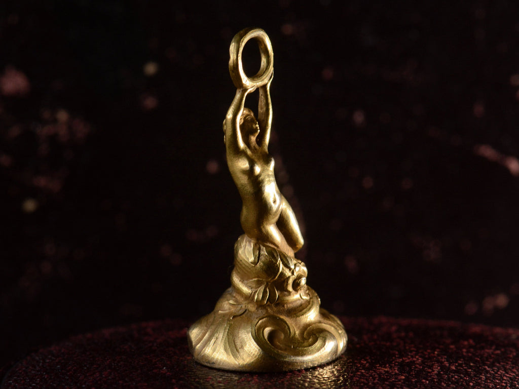 Left angled view of antique gold watch fob with a nude women resting on a base of vines, flowers and flowing water.  Shown on dark background.