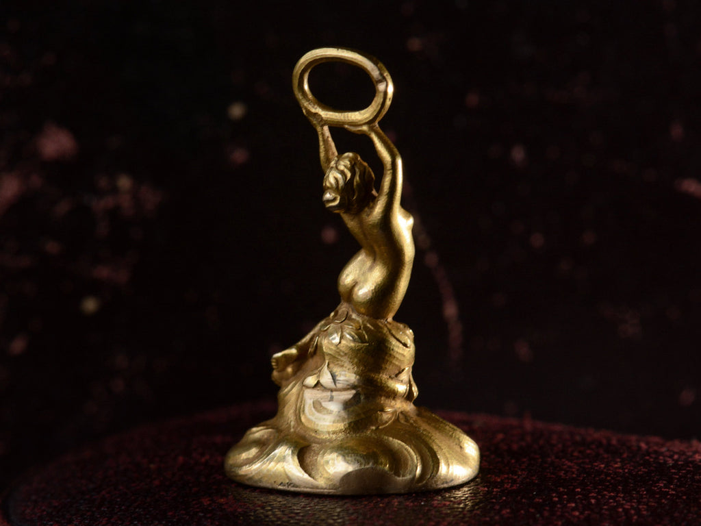 Back left view of antique gold watch fob with a nude women resting on a base of vines, flowers and flowing water.  Shown on dark background.