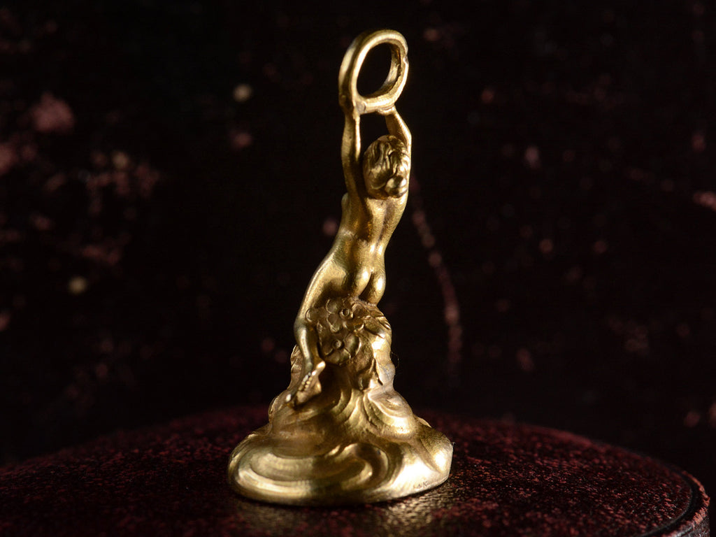 Back right view of antique gold watch fob with a nude women resting on a base of vines, flowers and flowing water.  Shown on dark background.