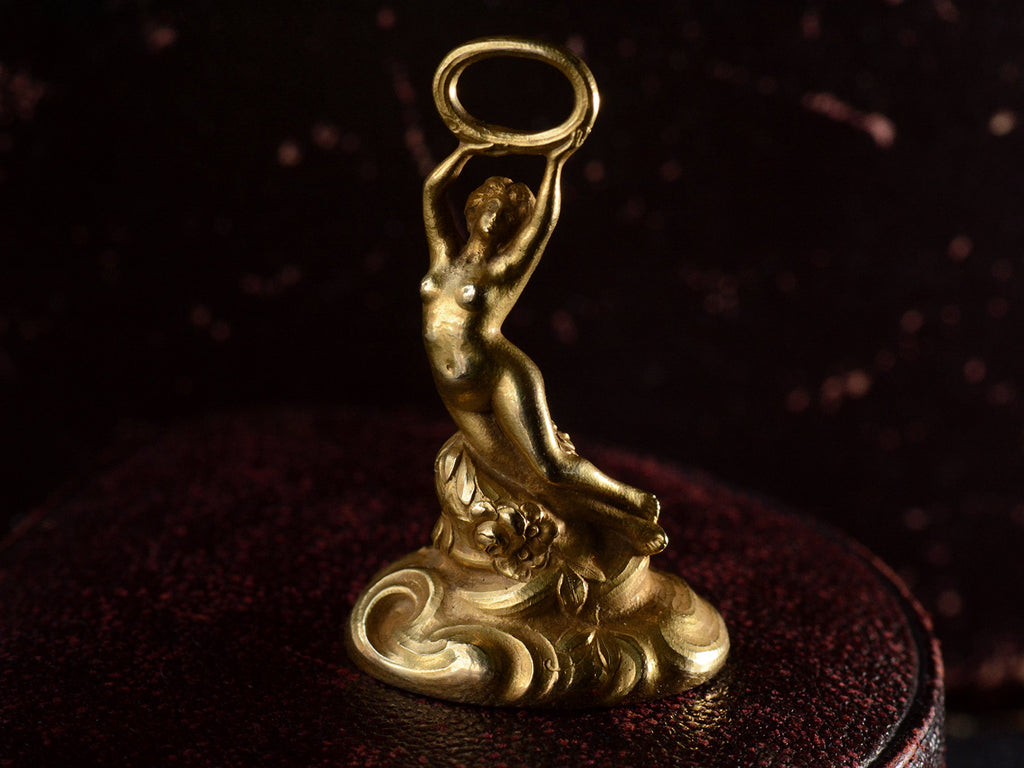 Elevated front view of antique gold watch fob with a nude women resting on a base of vines, flowers and flowing water.  Shown on dark background.