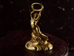 thumbnail of Elevated front view of antique gold watch fob with a nude women resting on a base of vines, flowers and flowing water.  Shown on dark background.