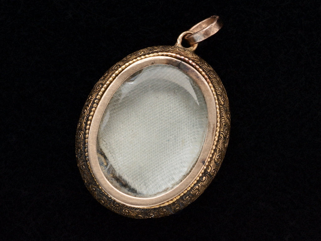 Backside view of oval antique gold locket with shown glass compartment with gray silk beneath it, shown on dark background.