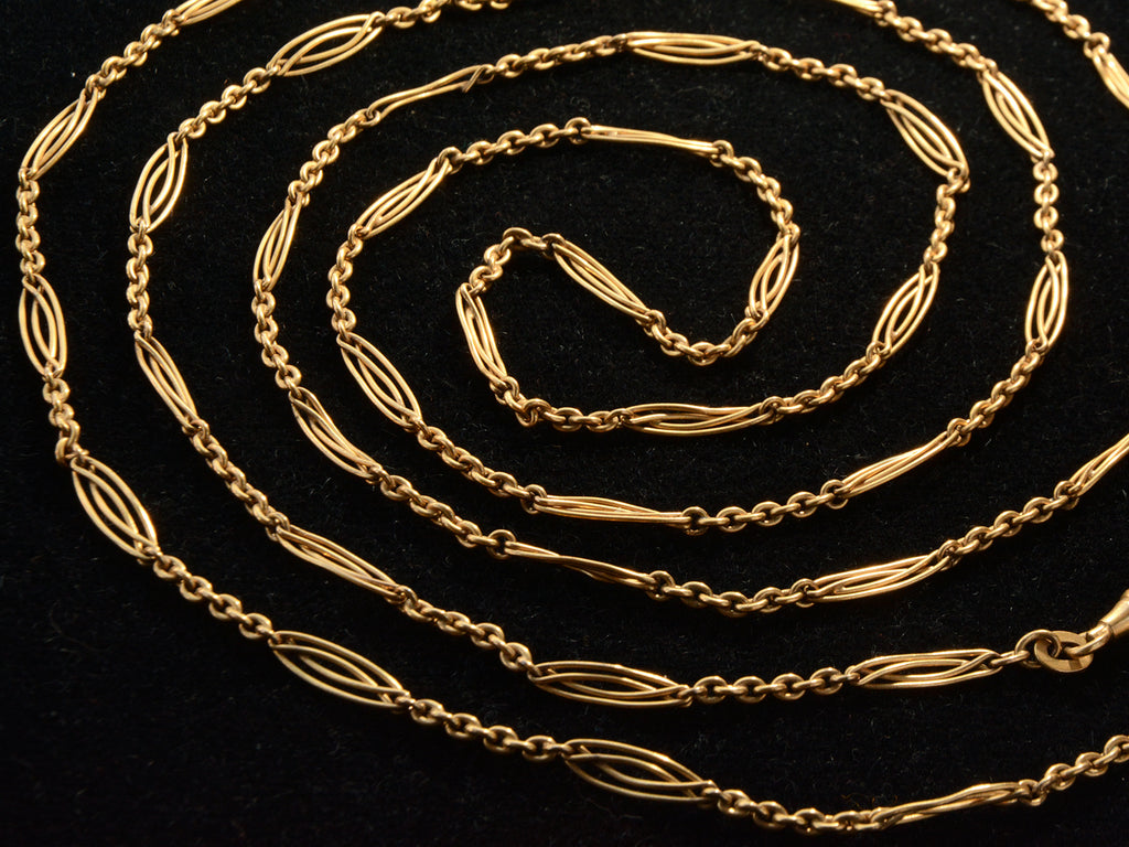 A close up view of long filigreed yellow gold plated c1890 chain, shown spiraled on a black background.