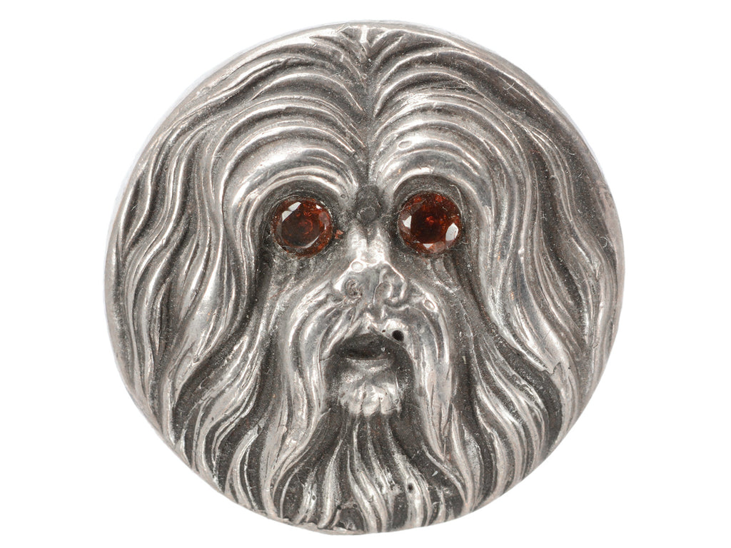 Round silver brooch with the face of a maltese dog with garnet eys, shown on white background.
