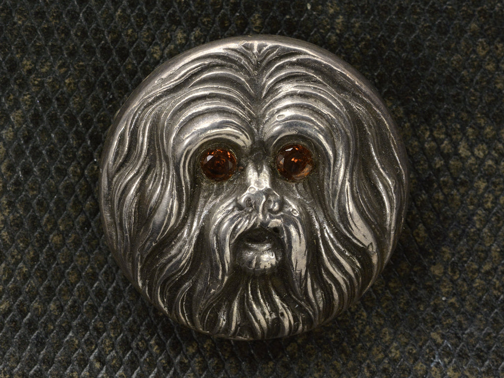 Round silver brooch with the face of a maltese dog with garnet eys, shown on dark textured background.