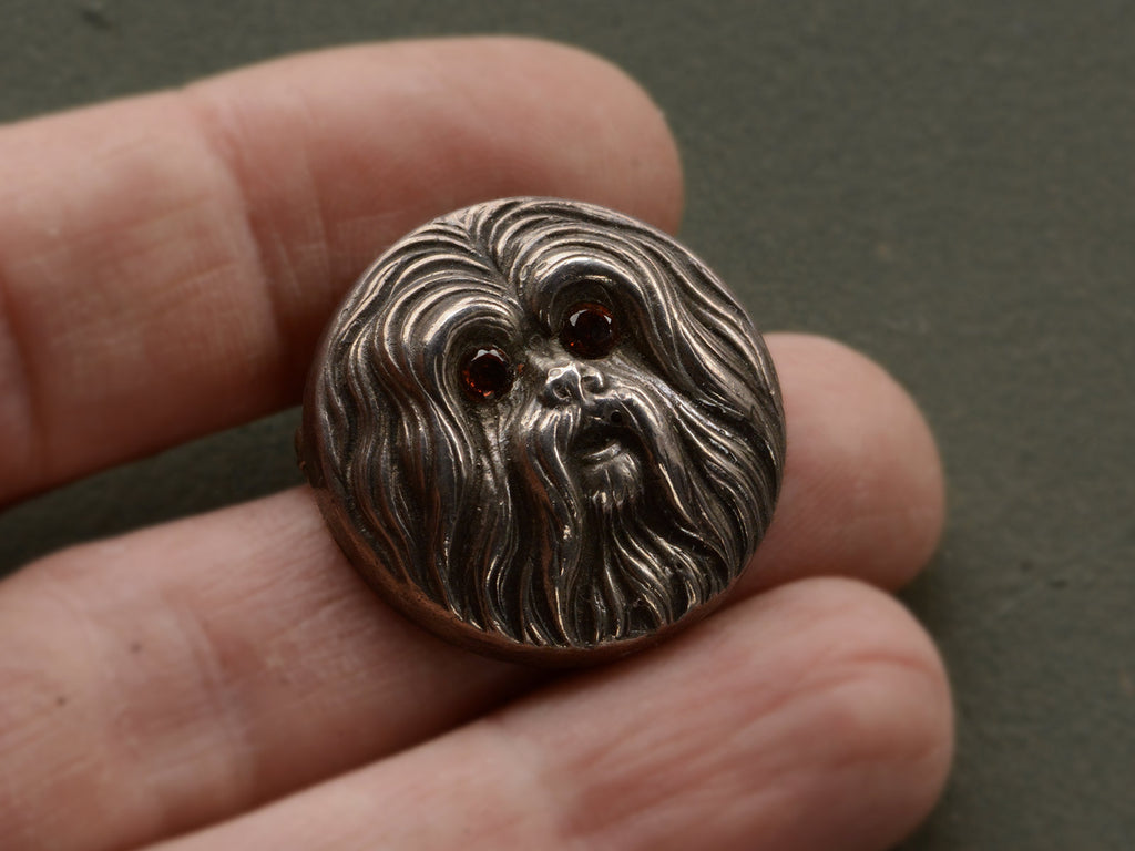 Round silver brooch with the face of a maltese dog with garnet eys, shown held on hand.