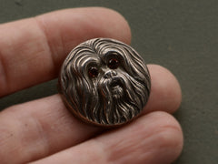thumbnail of Round silver brooch with the face of a maltese dog with garnet eys, shown held on hand.