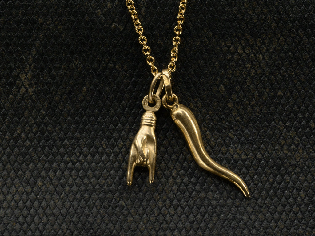 Detail of vintage 18K Italian charms, one of a hand gesturing the sign of the horns, the other of a cornicello horn, hanging together of cable chain. Shown on dark textured background.