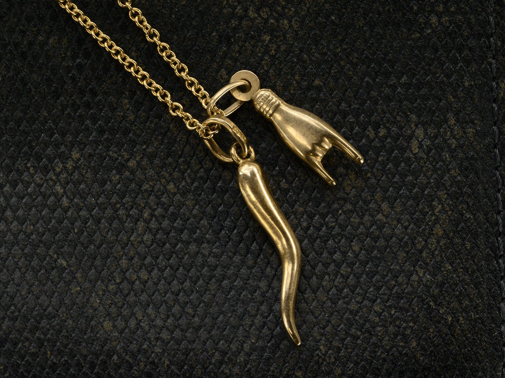Detail of alternate side of vintage 18K Italian charms, one of a hand gesturing the sign of the horns, the other of a cornicello horn, hanging together of cable chain. Shown on dark textured background.