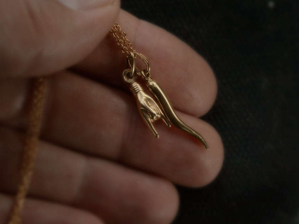Detail of a hand holding vintage 18K Italian charms, one of a hand gesturing the sign of the horns, the other of a cornicello horn, hanging together of cable chain. 