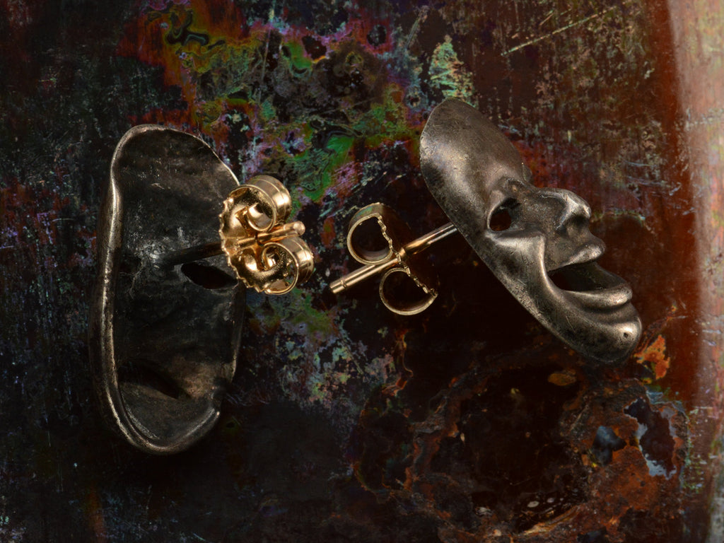 Side and back view of pair of oxidized silver theatrical mask stud earrings depicting tragedy and comedy, showing yellow gold fittings. On dark colorful background.