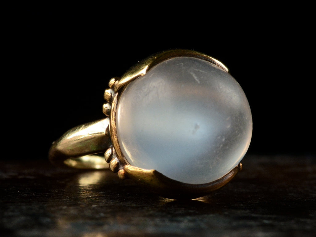Left angled view of vintage gold ring with silvery blue moonstone orb, shown on dark background.
