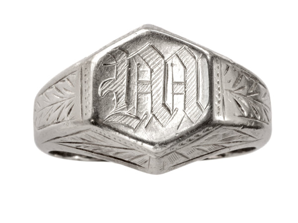 Front view of white gold Art Deco signet ring with a hexagonal top carved with a gothic script letter "M" and carved decorations on the sides, shown on white background.