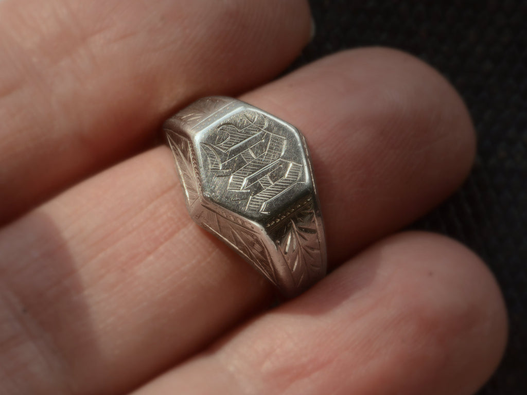 Front view of white gold Art Deco signet ring with a hexagonal top carved with a gothic script letter "M" and carved decorations on the sides, shown on finger