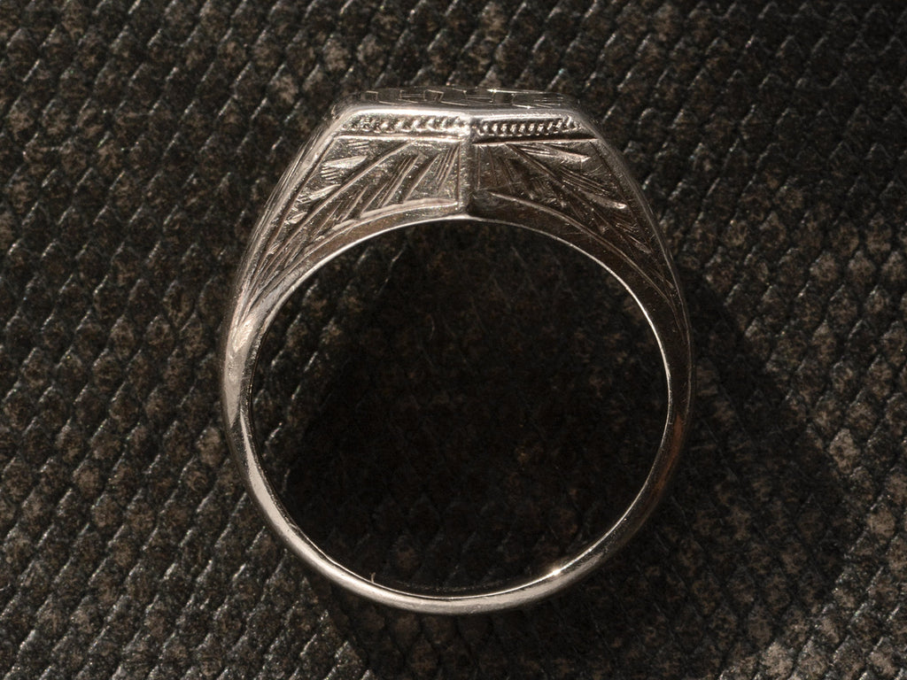 Side profile view of white gold Art Deco signet ring with carved decorations on the sides, shown on dark textured background.