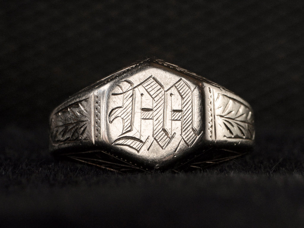Front view of white gold Art Deco signet ring with a hexagonal top carved with a gothic script letter "M" and carved decorations on the sides, shown on black background.