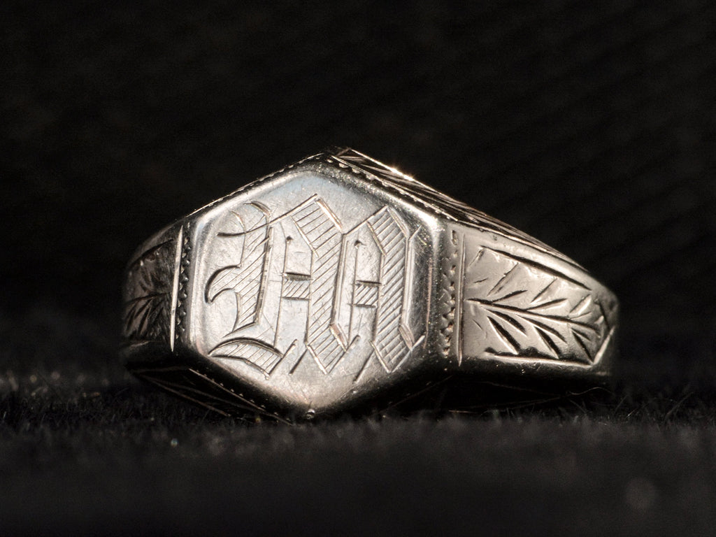Angled view of white gold Art Deco signet ring with a hexagonal top carved with a gothic script letter "M" and carved decorations on the sides, shown on dark background.