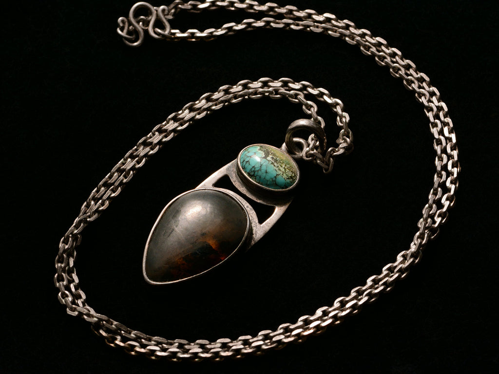 Angled view of silver pendant with oval turquoise and pear shaped tortoiseshell, on long chain, on black background.