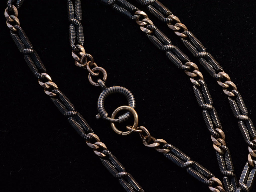 Close up of an antique chain of alternating silver and black striped links and gold filled links, focusing on clasp. Shown on black.