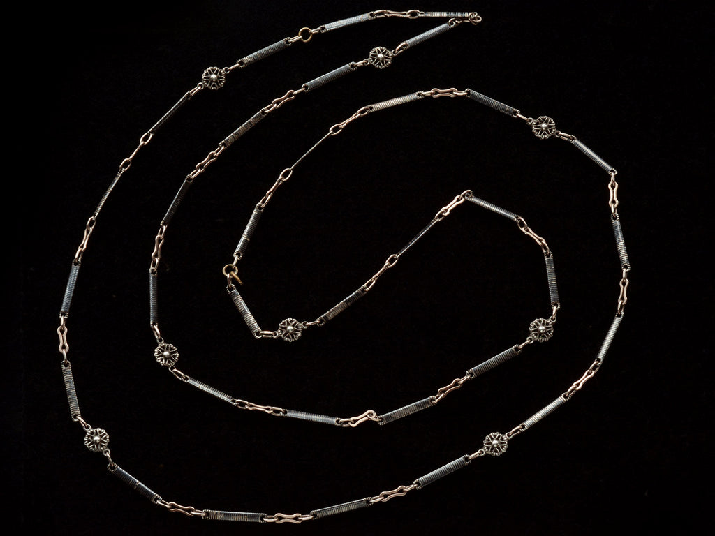 A 19th century long chain with alternating niello striped bar links, gold filled links, and silver rosettes, shown spiraled on black background.