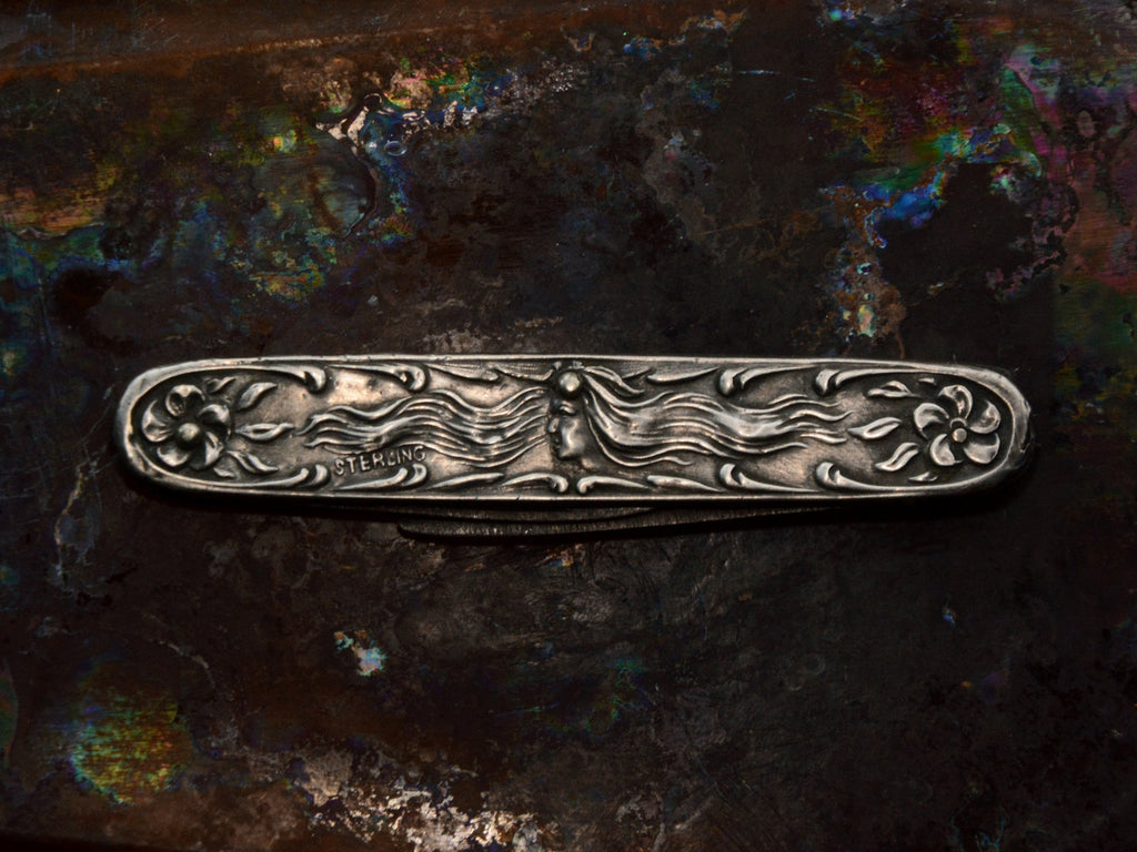 Art Nouveau silver pocket knife with imagery of a woman and flowers, shown on dark iridescent background.