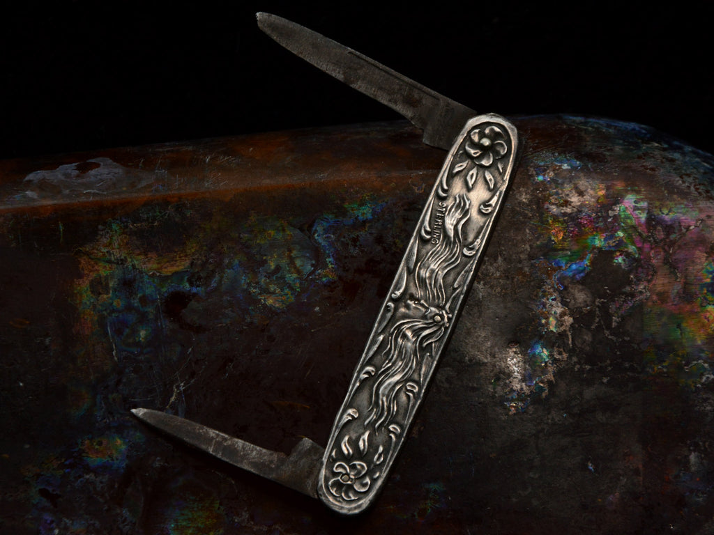 Art Nouveau silver pocket knife with imagery of a woman and flowers, shown with blades open on dark iridescent background.