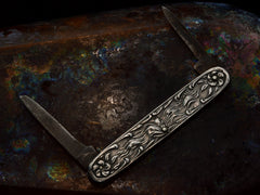 thumbnail of Reverse side of Art Nouveau silver pocket knife with imagery of a woman and flowers, shown with blades open on dark iridescent background.
