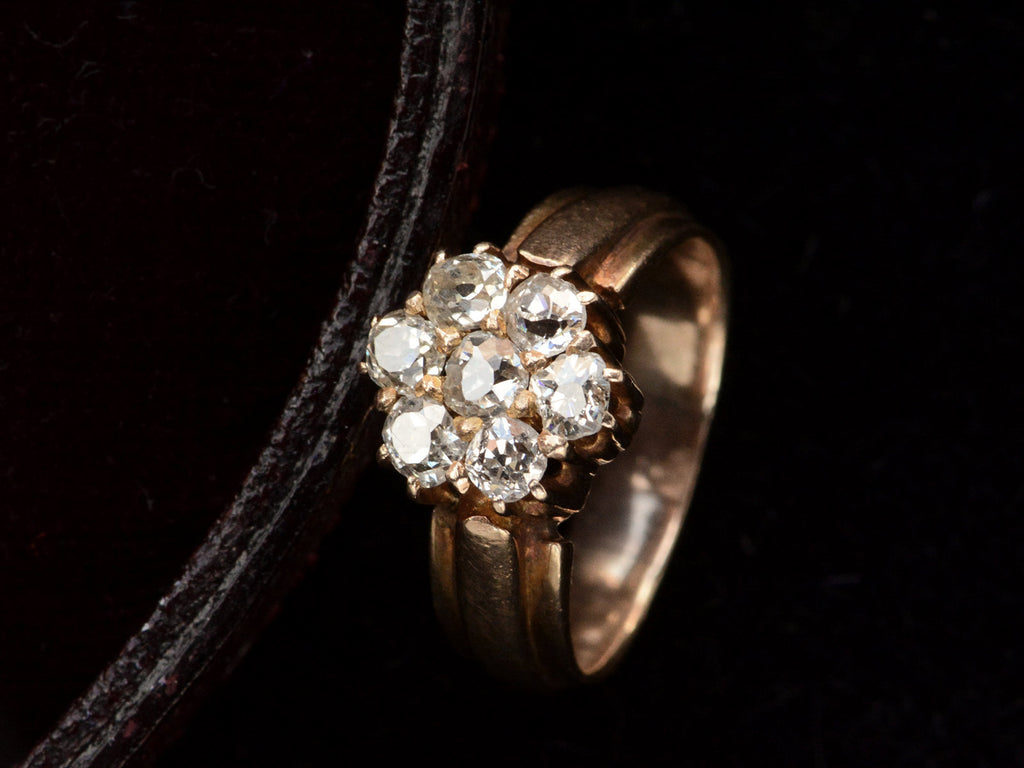 Angled view of antique rosy-yellow gold ring with cluster of old mine cut diamonds, on black background.