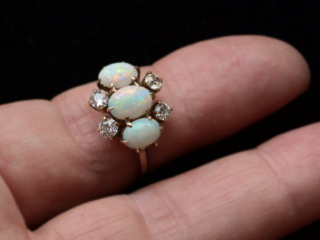 A vintage gold ring featuring three oval white opal stones and four round diamonds, set in gold shown on finger.