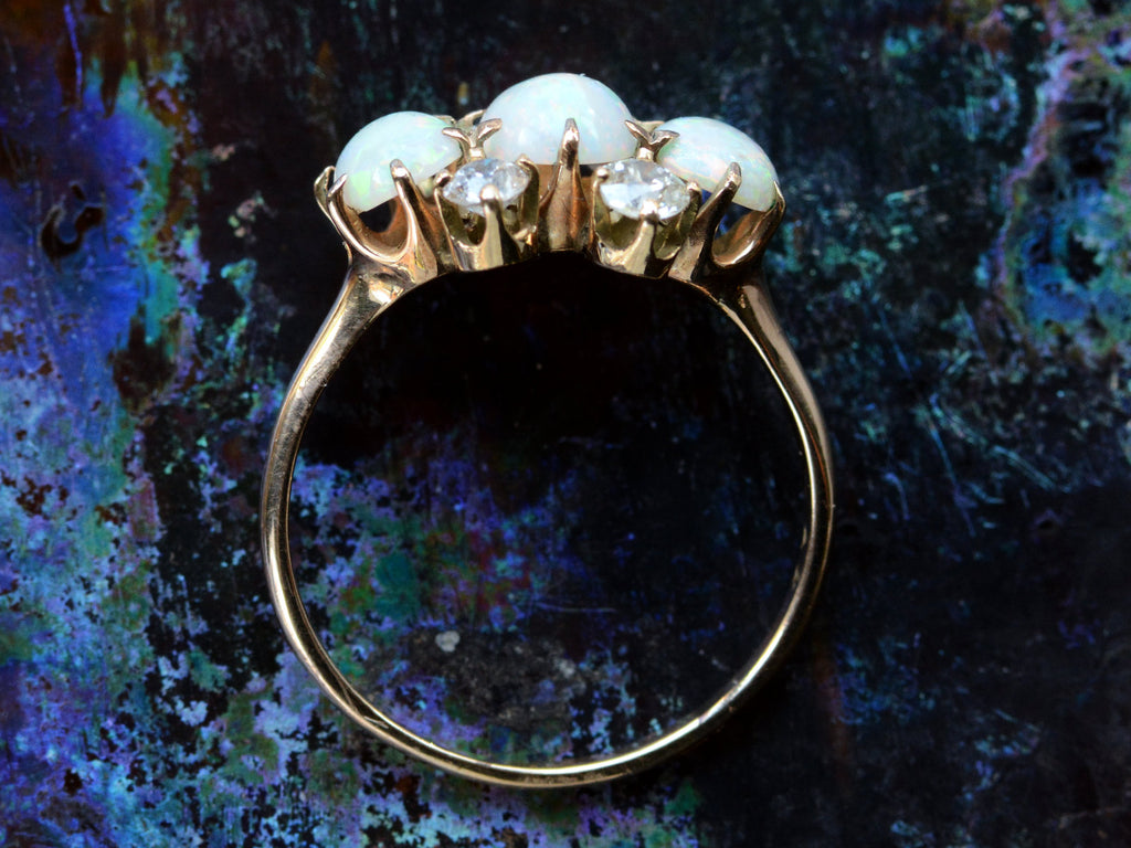 A side profile of a vintage gold ring featuring three oval white opal stones and four round diamonds, set in gold against a dark iridescent background.