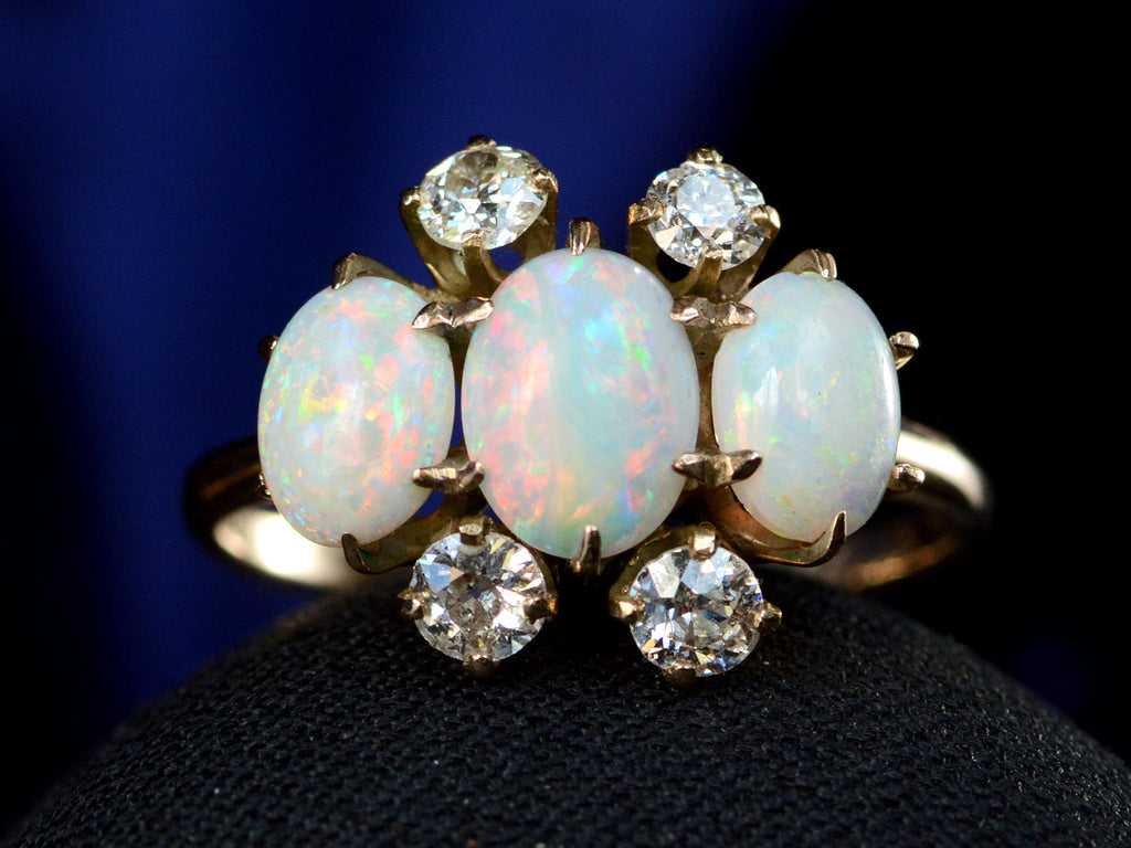 A gold ring featuring three oval white opal stones and four round diamonds, displayed on a black surface against a dark blue background.