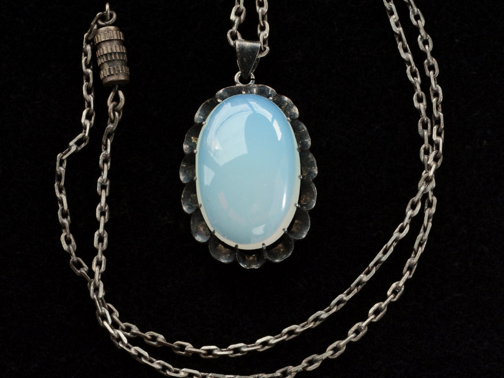 Detail of a vintage oxidized silver necklace with an oval opaline glass pendant, shown with chain clasp, on black background.