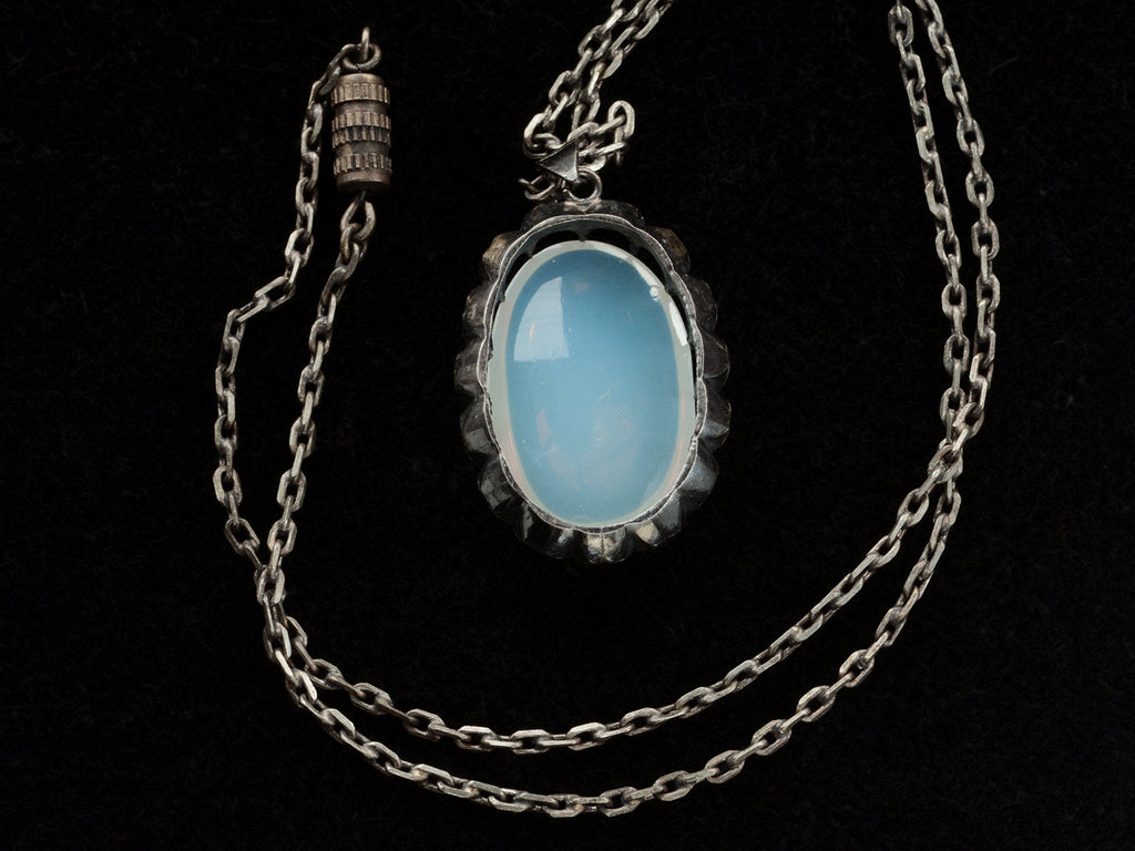 Reverse side detail of a vintage oxidized silver necklace with an oval opaline glass pendant, shown with chain clasp, on black background.