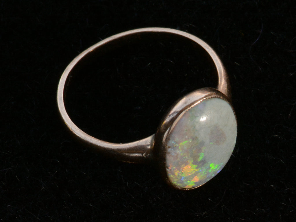 Tilted view of c1920 oval opal ring in rosy 10K gold, shown on black background.