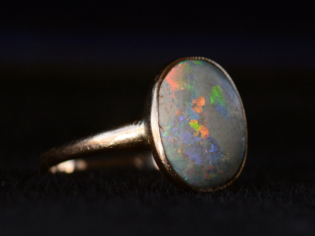 Left angled view of c1920 oval opal ring in rosy 10K gold, shown on black background.