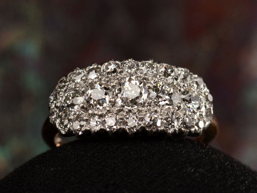 Front view of antique c1900 ring with an oval cluster of round old European cut diamonds set in platinum-topped 14K yellow gold, shown on dark background.