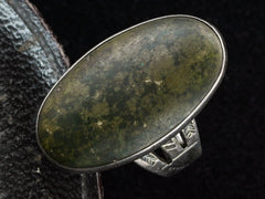 thumbnail of Angled view of a vintage Navajo ring with a large dark olive green cabochon bezel set in silver with a decorative stamped shank. Shown on black background.