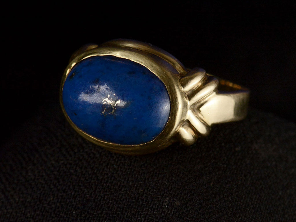 Angled view of vintage c1980 ring with an oval blue lapis lazuli set in 18K yellow gold, shown on dark background.