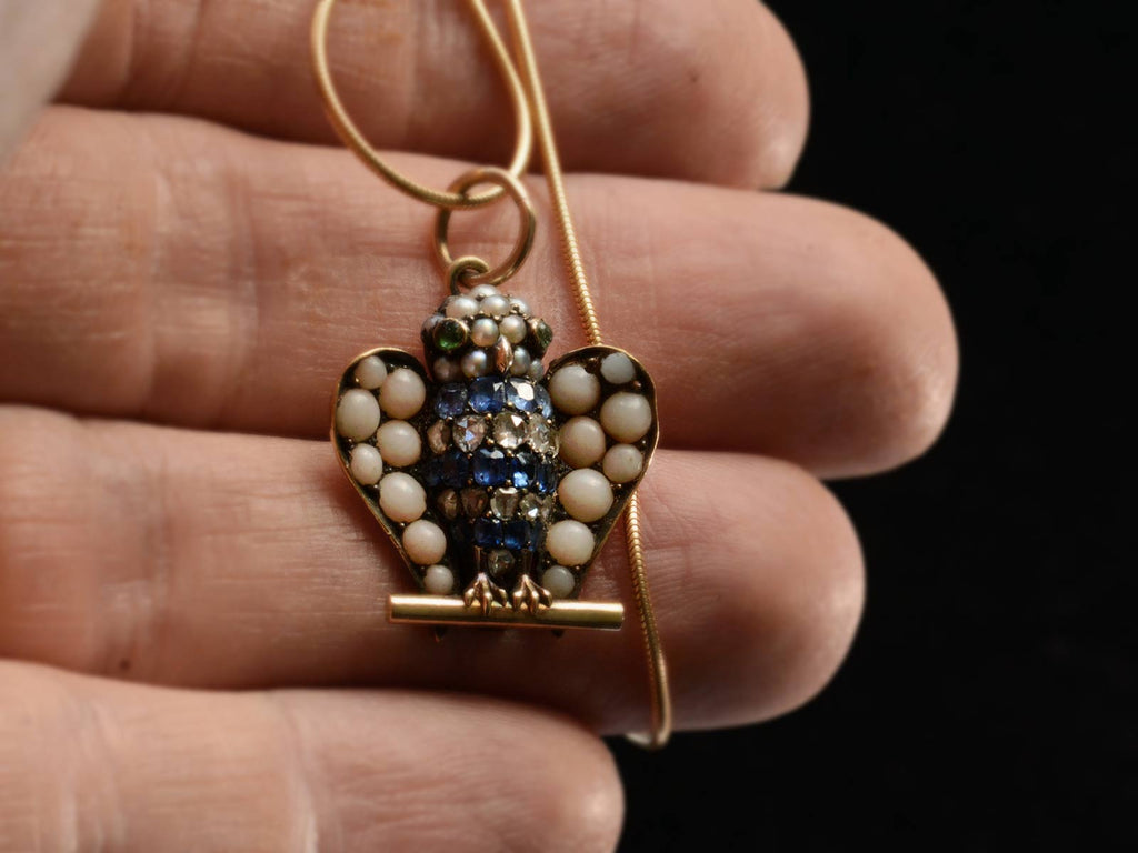 A owl pendant set with pale pink coral, sapphires, diamonds, pearls, and emeralds (shown on hand for scale)