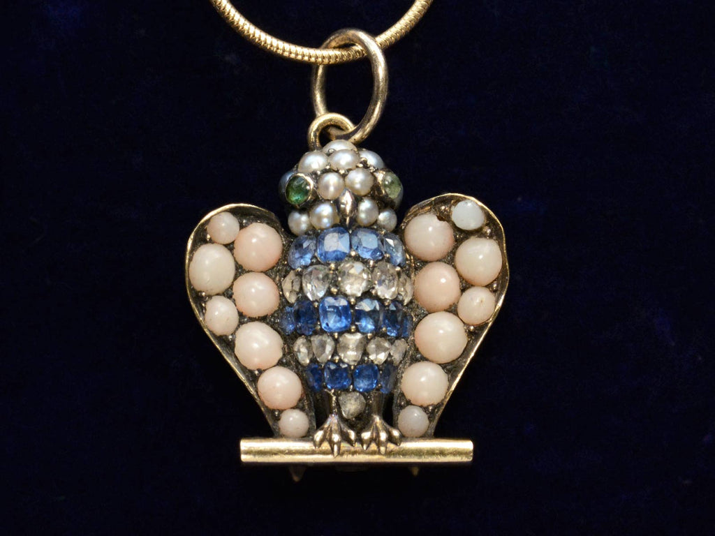 A owl pendant set with pale pink coral, sapphires, diamonds, pearls, and emeralds (shown on dark purple background)