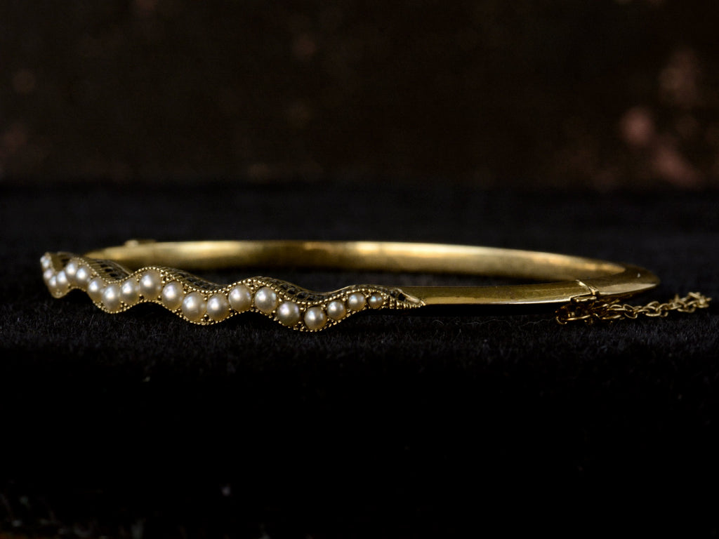 Right angled view of thin gold c1900 Art Nouveau bracelet with a wavy line of pearls. Shown on dark background.