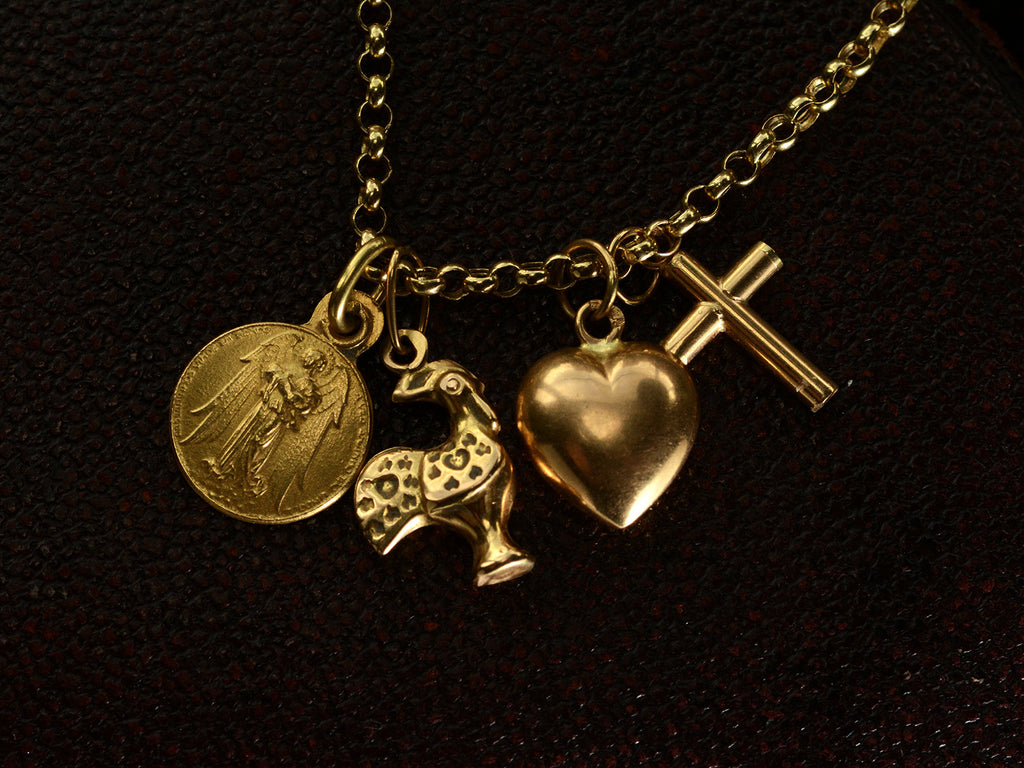 Detail of vintage 19k yellow gold Portuguese charm necklace with four charms: a devotional medallion of Christ, a rooster, a heart, and a cross. Shown against dark texutred background.