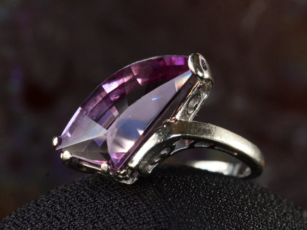 Angled view white gold ring with wing shaped faceted purple stone, on dark background.