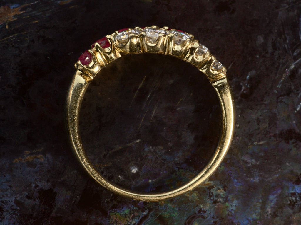 Side profile of c1970 Yellow Gold Ring with rows of pink rubies and white diamonds curling around each other. On dark background.