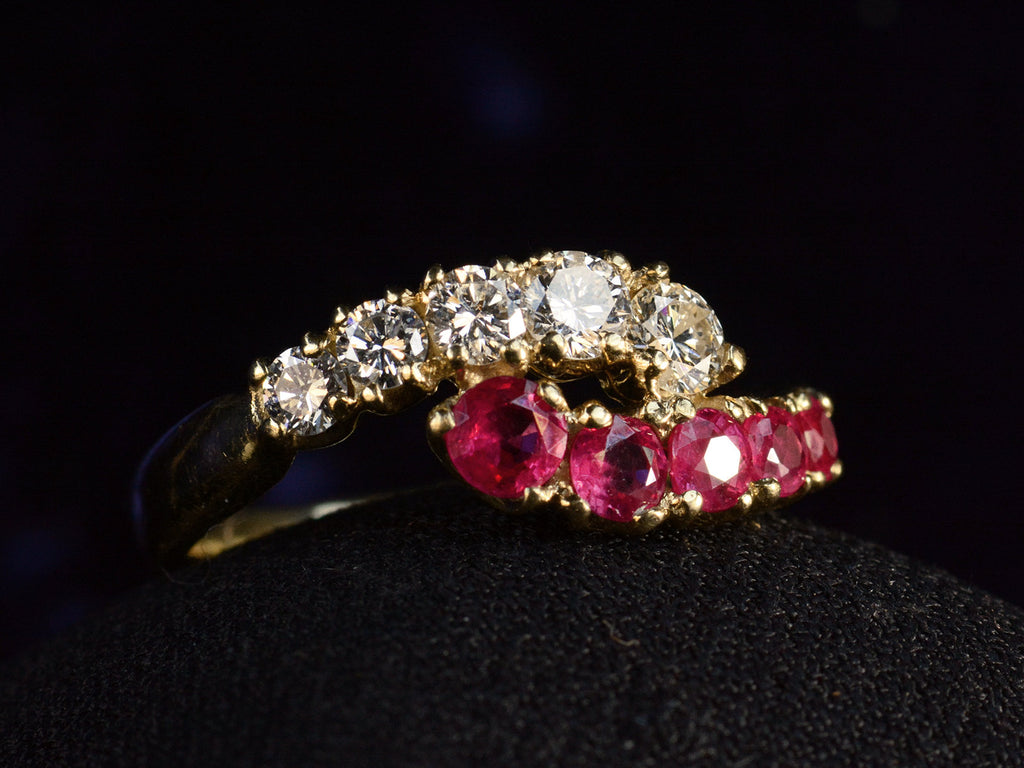 Angled view of c1970 Yellow Gold Ring with rows of pink rubies and white diamonds curling around each other. On dark background.