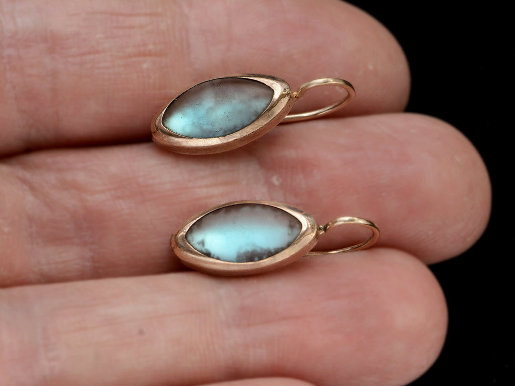 Pair of gold earrings with marquise shaped iridescent pale blue saphiret cabochons, shown on hand.