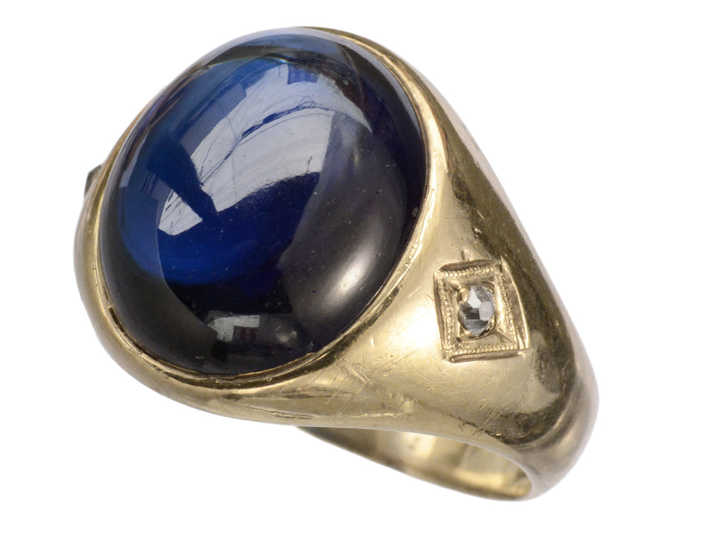 Wide gold antique ring with a large oval blue cabochon and diamonds on the sides, shown on white background.