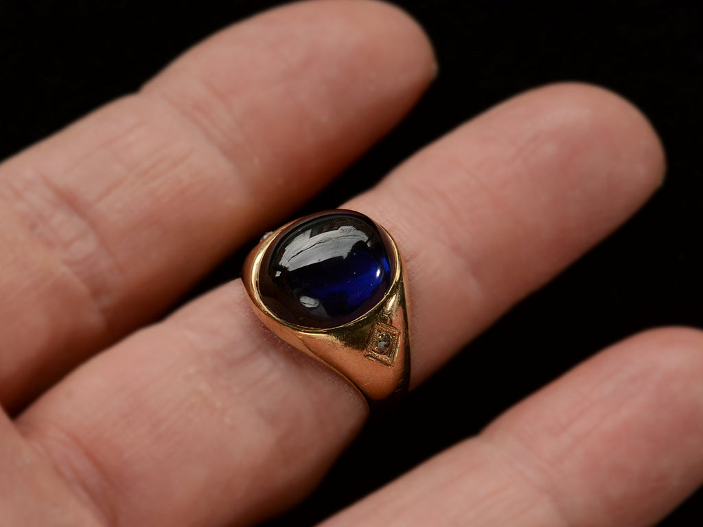 Wide gold antique ring with a large oval blue cabochon and diamonds on the sides, shown on finger.