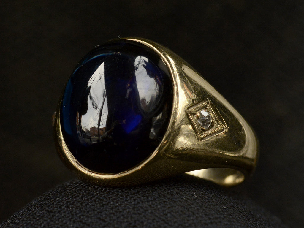 Angled front view of wide gold antique ring with a large oval blue cabochon and diamonds on the sides, shown on dark background.