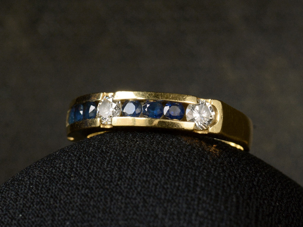 Angled view of a vintage yellow gold ring with three diamonds interspersed with blue sapphires. Shown on dark background.